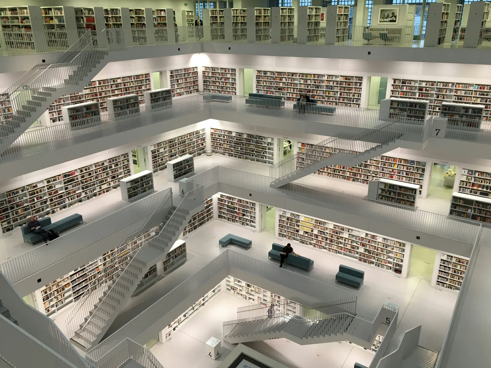 Bibliothèque moderne sur plusieurs étages avec escaliers blancs. Plusieurs personnes lisent ou se détendent sur des bancs. Atmosphère calme.