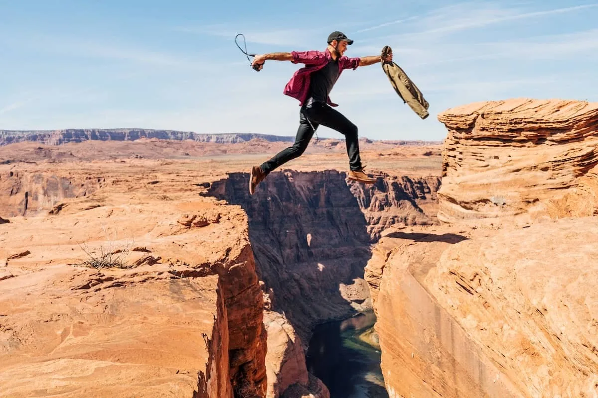 Un homme saute entre deux falaises du Grand Canyon, portant une chemise rouge et un sac. Ciel bleu clair et paysage désertique.