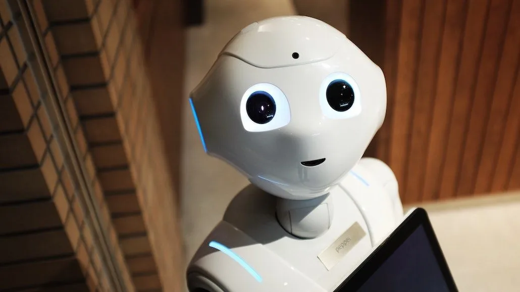 Un robot blanc avec des yeux lumineux se tient dans un intérieur en bois et briques. Il semble amical et curieux.