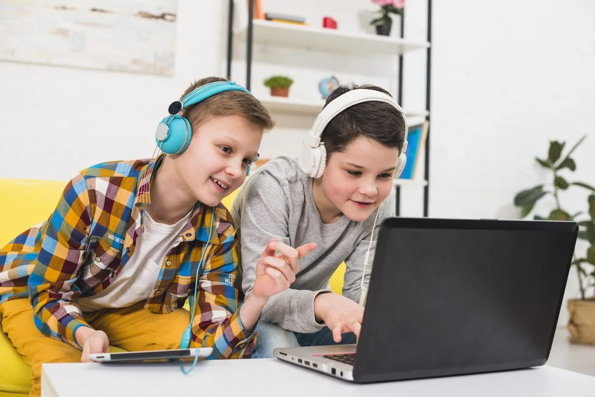 Deux garçons souriants, casque audio, sur un ordinateur portable. L'un porte une chemise à carreaux colorée. Intérieur lumineux.