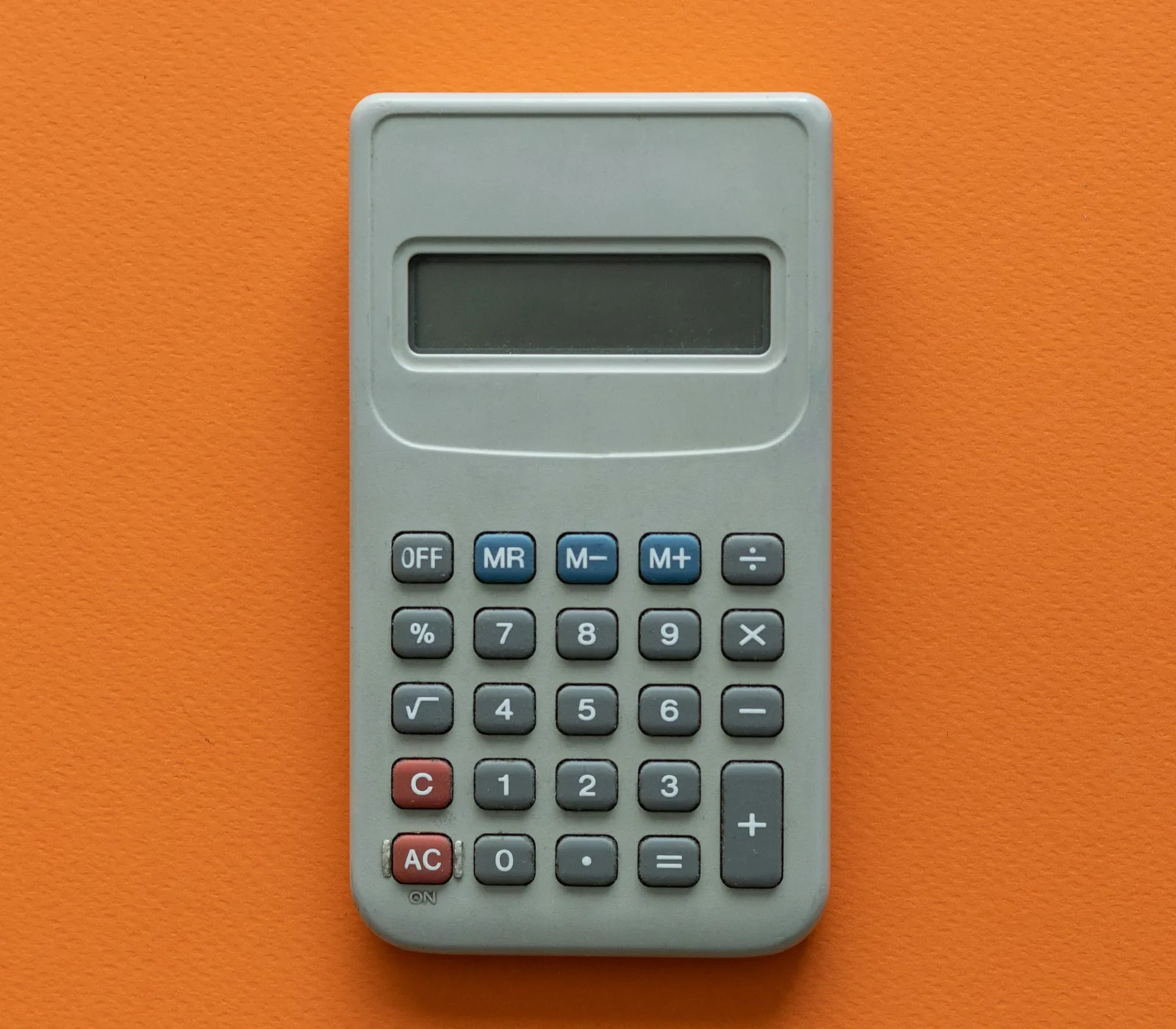 Calculatrice grise sur fond orange vif. Les touches sont rouges, bleues et grises avec des symboles et chiffres. Mood neutre.