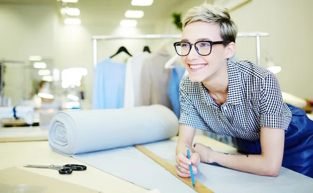 Une femme souriante avec des lunettes travaille dans un atelier de couture, mesurant du tissu bleu. Vêtements suspendus en arrière-plan.