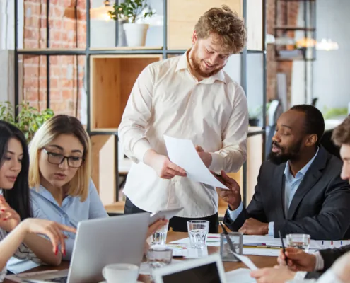 Un groupe de collègues divers discute autour d'une table dans un bureau moderne. Un homme souriant remet des documents, ambiance collaborative.
