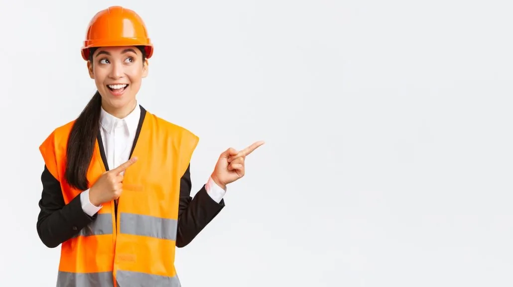Femme en casque et gilet orange, sourit et montre des deux mains à droite. Fond blanc, ambiance joyeuse et professionnelle.