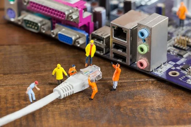Des figurines miniatures habillées en ouvriers manipulent un câble Ethernet géant sur une carte mère colorée posée sur une table en bois.